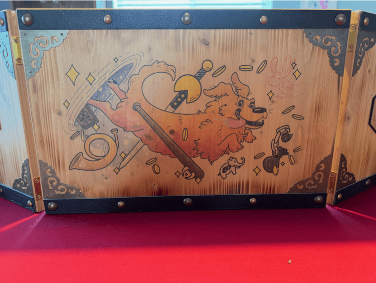 A wooden GM screen displayed on a red table. In the middle of the screen is a hand-drawn picture of a cartoonish golden retriever emerging from a purple portal. The dog has a huge dopey smile and his tongue is hanging out. He is surrounded by coins, sparkles, and items from the Deck of Silly Things including a wallet with a chain, an elephant statue, a looping horn, a baseball bat with fangs, bunny slippers, and a sword with pacman on the hilt
